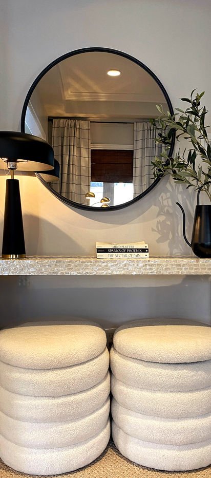 A round black mirror above a shelf, a black lamp with a brass band, and two boucle stools.