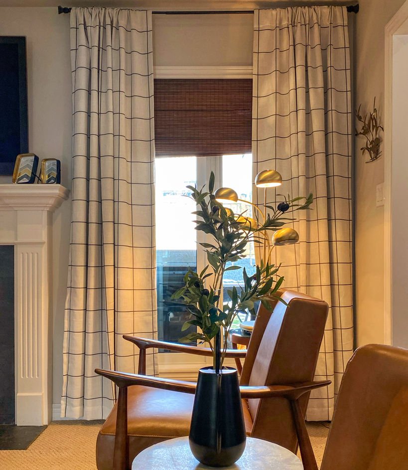 Tall windows with checked drapes and a woven blind, olive branches in a black vase, and two tan leather mid-century chairs.