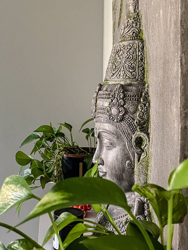 A carved stone figure beside trailing pothos and monstera leaves against a sheer curtain.
