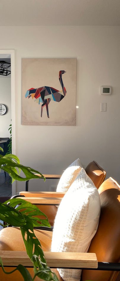 A colourful painterly ostrich print above a tan leather sofa with cream cushions, beside a monstera.